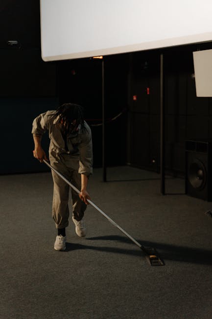 A woman using a yellow vacuum cleaner to deep clean an ornate, patterned area rug with floral motifs and a muted color palette, situated on a wooden floor in a living room. The room features a leather