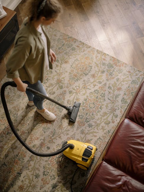A living room scene showing a clean wooden floor illuminated by natural light from windows. In the foreground, there is a vacuum cleaner with a transparent dust container, placed next to a neatly arra